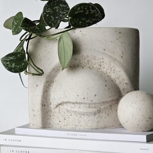 May include: A white ceramic planter with a textured surface and a raised eye design. A green plant with variegated leaves is in the planter. The planter is sitting on top of a stack of three magazines. The top magazine is titled "GUIDED LONDON", the middle magazine is titled "IN CLOVER", and the bottom magazine is titled "CEREAL 09".