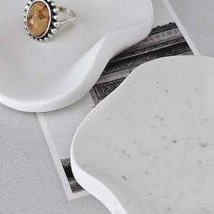 Op de afbeelding: Een zilveren ring met een ovale, amberkleurige steen ligt op een witte, organisch gevormde mini-schaal. De schaal maakt deel uit van een handgemaakte decoratieset. De tekst "ALBA ORGANIC MINI DISH" is zichtbaar.