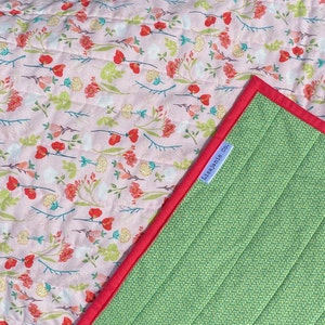 May include: A pink and green reversible quilt with a floral print on one side and a green quilted pattern on the other. The quilt has a red binding and a white label that says "Lila Jane Co."