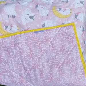 May include: Pink and white quilted baby blanket with a yellow border. The blanket features a repeating pattern of white sheep and yellow moons. The back of the blanket is pink with white numbers printed on it. The blanket has a tag that says "Lizajanie Co."