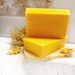 May include: Two bars of bright yellow soap stacked on top of each other. The soap is on a white surface with a light brown textured surface underneath.