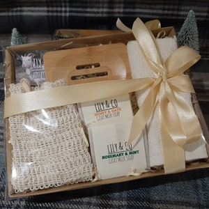 May include: A gift basket containing a white face cloth, a wooden soap dish, a natural loofah, and two bars of goat milk soap. The soap bars are labelled "LILY & CO HONEY & OATS GOAT MILK SOAP" and "LILY & CO ROSEMARY & MINT GOAT MILK SOAP". The basket is tied with a gold ribbon.
