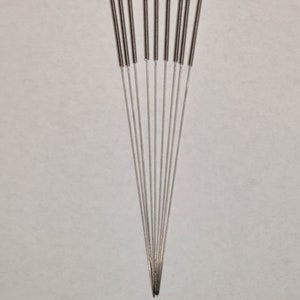 Könnte beinhalten: Nahaufnahme eines Fächers von Akupunkturnadeln. Jede Nadel hat einen dünnen, silbernen Schaft mit einem gewendelten Griff. Die Nadeln sind fächerförmig nach unten auf einem schlichten, hellen Hintergrund angeordnet.