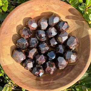 May include: A bowl filled with a collection of dark red garnet tumbled stones. The stones are smooth and polished, with a variety of shapes and sizes.
