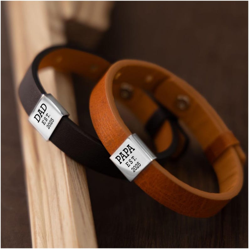 Op de afbeelding: Twee leren armbanden, &eacute;&eacute;n bruin en &eacute;&eacute;n lichtbruin, met zilveren sluitingen. De sluiting van de bruine armband leest "DAD EST. 2025", en die van de lichtbruine armband "PAPA EST. 2025". De armbanden liggen op een houten oppervlak.