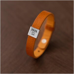 Op de afbeelding: Een bruin lederen armband met een zilveren rechthoekige sluiting. De sluiting is gegraveerd met het woord "PAPA" boven "EST. 2025". De armband is een enkele band met een gespsluiting. De achtergrond is een donkerbruin houten oppervlak.