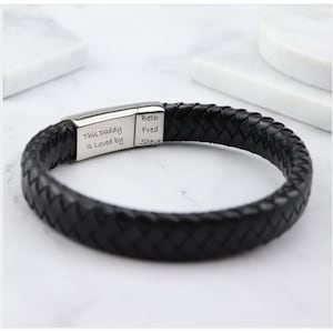 Puede incluir: Pulsera de cuero trenzado negro con cierre plateado. El cierre está grabado con las palabras "This Daddy Is Loved by" y los nombres Beth, Fred y Steve. Un accesorio elegante.