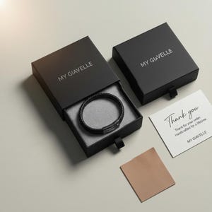 May include: A black leather bracelet with a braided design is displayed in an open black box. The box has the text "MY GIAVELLE" on the lid. A thank you card and a small tan cloth are also visible.