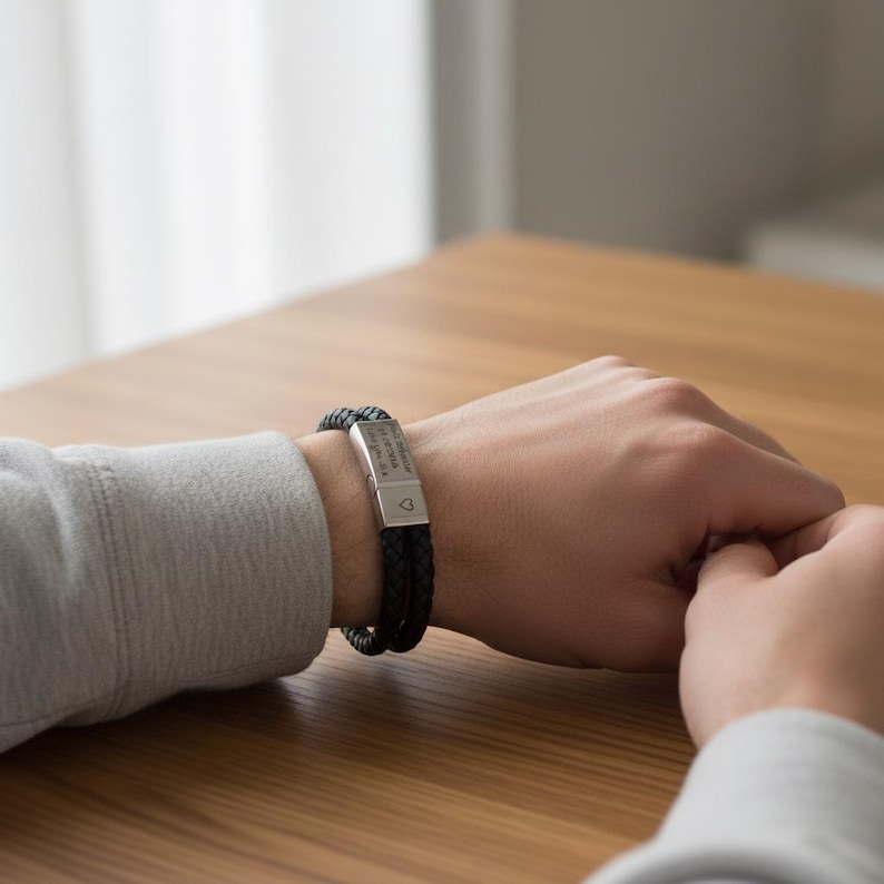 May include: Black braided leather bracelet with a silver clasp. The clasp has engraved text and a small heart symbol. The bracelet is worn on a wrist, with the person's hands resting on a wooden surface.