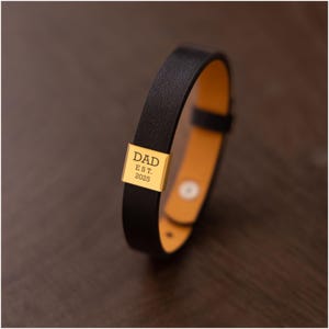 Op de afbeelding: Zwarte leren armband met een goudkleurige rechthoekige bedel. De bedel is gegraveerd met het woord "DAD" boven "EST. 2025". De armband heeft een lichtbruine binnenkant en een eenvoudige gespsluiting. Een stijlvol accessoire.