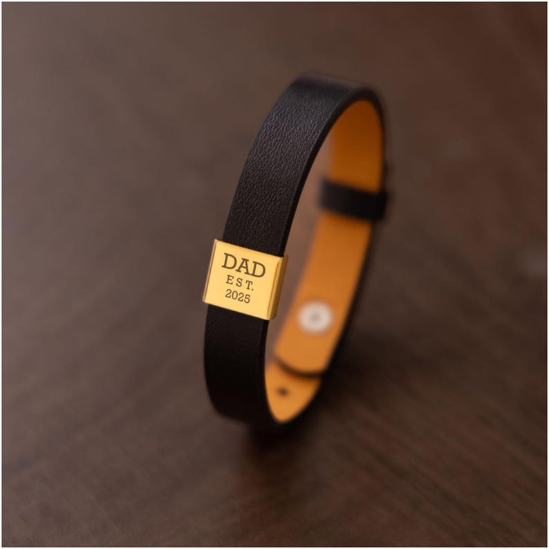 Op de afbeelding: Zwarte leren armband met een goudkleurige rechthoekige bedel. De bedel is gegraveerd met het woord "DAD" boven "EST. 2025". De armband heeft een lichtbruine binnenkant en een eenvoudige gespsluiting. Een stijlvol accessoire.