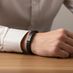 May include: Black braided leather bracelet with a rectangular metal clasp. The clasp is engraved with the words "Happy 21st Birthday" and a heart symbol. The bracelet is worn on a wrist.