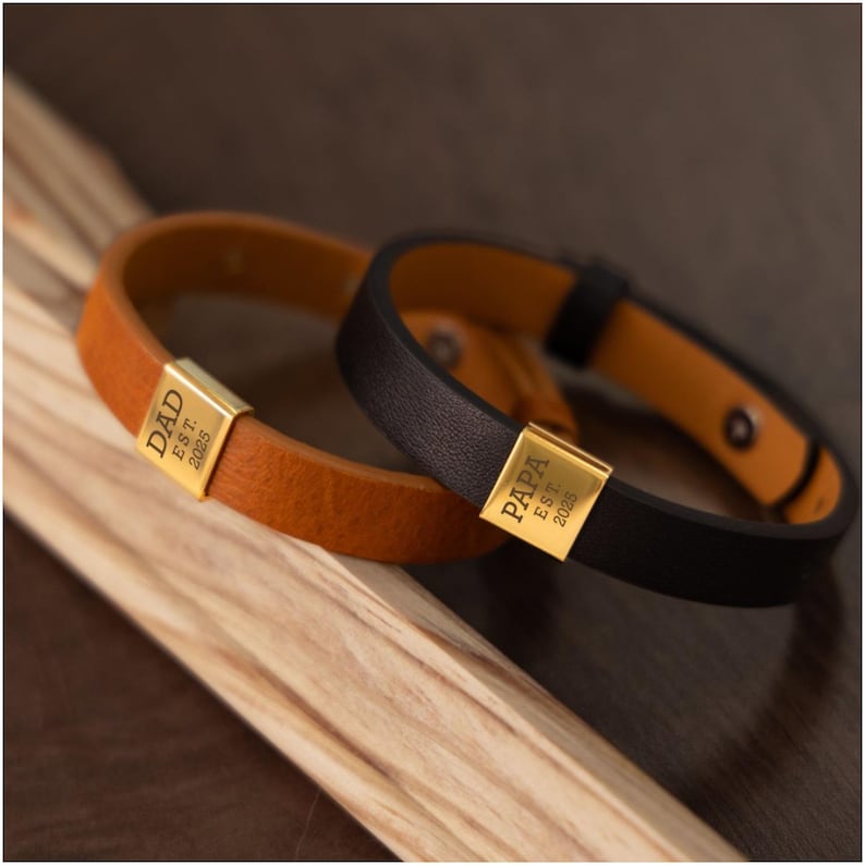 Op de afbeelding: Twee leren armbanden, &eacute;&eacute;n bruin en &eacute;&eacute;n zwart, met goudkleurige rechthoekige sluitingen. De bruine armband leest "DAD EST. 2025" en de zwarte armband leest "PAPA EST. 2025". De armbanden liggen op een houten oppervlak.