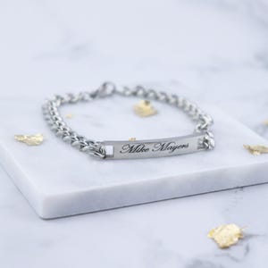 Puede incluir: Una pulsera de cadena de eslabones plateada con una placa rectangular con el nombre "Mike Mayers" grabado. La pulsera se exhibe sobre una superficie de mármol blanco con detalles de hojas de oro.