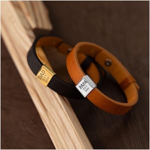 Op de afbeelding: Twee leren armbanden, &eacute;&eacute;n zwart en &eacute;&eacute;n bruin, met metalen sluitingen. De zwarte armband heeft een gouden sluiting met de gravure "DAD EST. 2025". De bruine armband heeft een zilveren sluiting met de gravure "PAPA EST. 2025".
