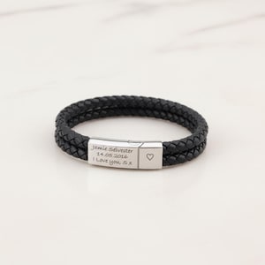 May include: Black braided leather bracelet with a silver clasp. The clasp is engraved with the text "Jamie Selvester 14.05.2016 I Love you, Sx" and a heart symbol. The bracelet is a stylish accessory.