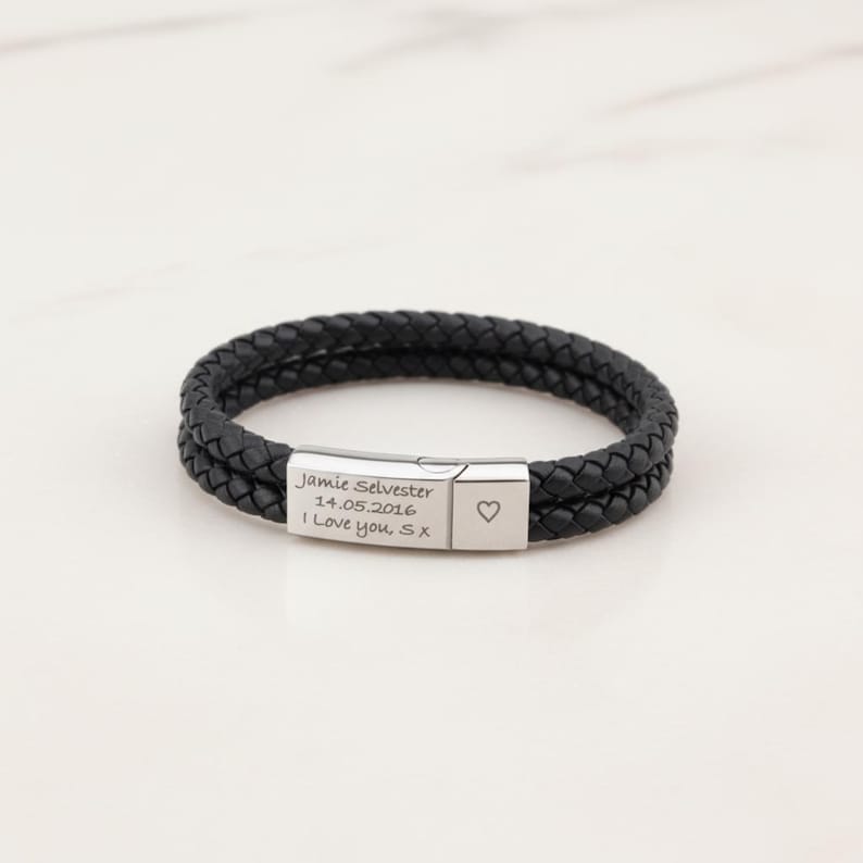 May include: Black braided leather bracelet with a silver clasp. The clasp is engraved with the text "Jamie Selvester 14.05.2016 I Love you, Sx" and a heart symbol. The bracelet is a stylish accessory.