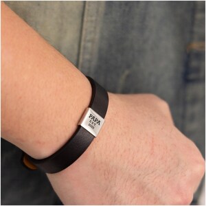 Op de afbeelding: Zwarte leren armband met een zilveren sluiting. De sluiting is gegraveerd met het woord "PAPA" boven "EST. 2025". De armband wordt om de pols gedragen.