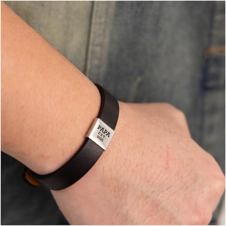 Op de afbeelding: Zwarte leren armband met een zilveren sluiting. De sluiting is gegraveerd met het woord "PAPA" boven "EST. 2025". De armband wordt om de pols gedragen.