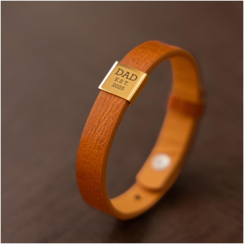 Op de afbeelding: Een bruin leren armband met een goudkleurige rechthoekige plaat. De plaat is gegraveerd met het woord "DAD" boven "EST. 2025". De armband heeft een eenvoudig ontwerp met een gespsluiting.