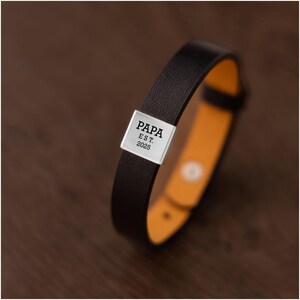Op de afbeelding: Een zwart leren armband met een zilveren rechthoekige plaat. De plaat is gegraveerd met het woord "PAPA" boven "EST. 2025". De binnenkant van de armband is felgeel-oranje.