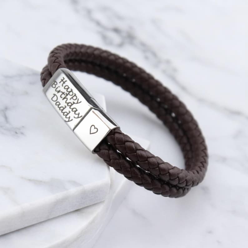 May include: A brown leather bracelet with a braided design. The silver clasp is engraved with "Happy Birthday Daddy" and a heart symbol. The bracelet is a stylish accessory.