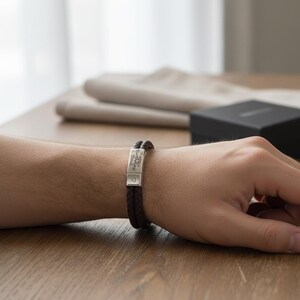 May include: A dark brown braided leather bracelet with a silver clasp. The clasp is engraved with the words "Happy Birthday Dad" and a small heart symbol. The bracelet is on a person's wrist.
