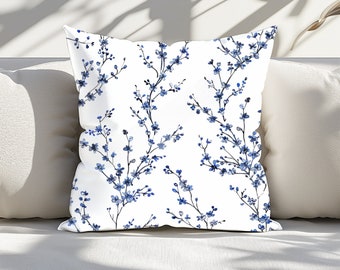 May include: Square white throw pillow with a repeating pattern of blue floral branches. The pillow is on a white surface, with shadows in the background. The floral design features small blue flowers and delicate stems.