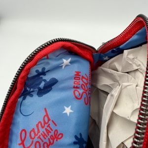 May include: A red zippered pouch with a light blue interior lining featuring a repeating pattern of white stars, dark blue silhouettes of Mickey Mouse, and the red text that reads Land That I Love From Sea To Shining Sea.