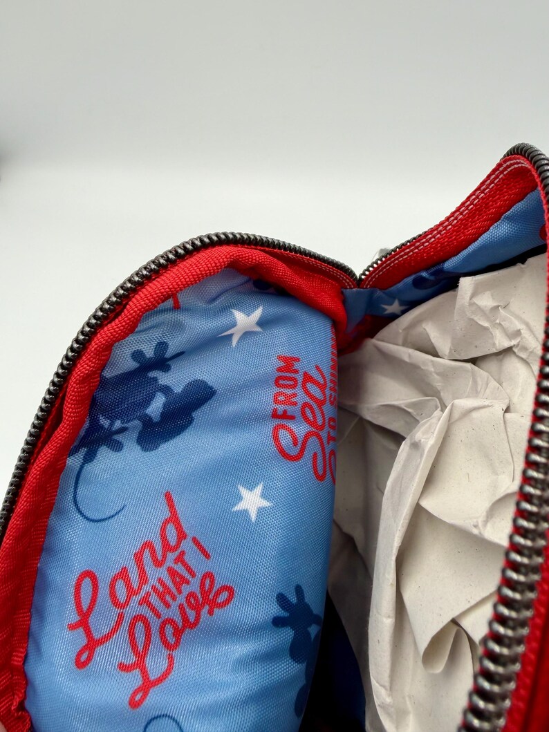 May include: A red zippered pouch with a light blue interior lining featuring a repeating pattern of white stars, dark blue silhouettes of Mickey Mouse, and the red text that reads Land That I Love From Sea To Shining Sea.