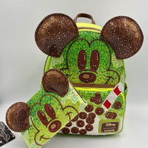 May include: A lime green backpack and matching wallet featuring a Mickey Mouse design. The backpack has brown ears and is embellished with sparkling accents. The wallet and backpack have a beverage design with a red and white straw.