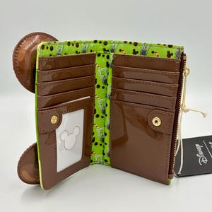 May include: Open brown wallet with a green interior featuring a pattern of drinks. The wallet has multiple card slots, a clear ID window with a Mickey Mouse outline, and a zipper closure. A brown circular element is attached to the wallet.