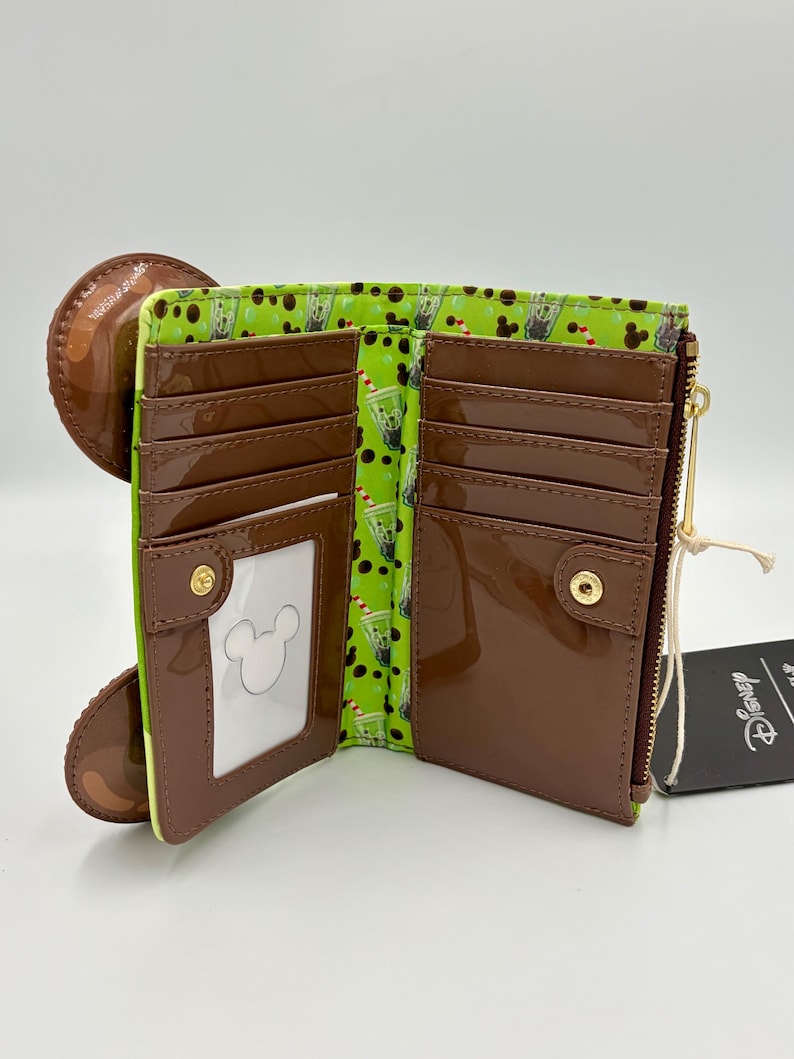 May include: Open brown wallet with a green interior featuring a pattern of drinks. The wallet has multiple card slots, a clear ID window with a Mickey Mouse outline, and a zipper closure. A brown circular element is attached to the wallet.