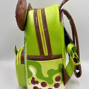 May include: A lime green backpack with brown accents and a yellow zipper. The backpack features a camouflage pattern with brown circles, and a brown ear-shaped embellishment. The straps are green and brown.