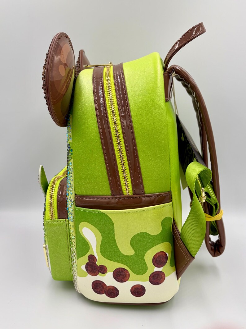 May include: A lime green backpack with brown accents and a yellow zipper. The backpack features a camouflage pattern with brown circles, and a brown ear-shaped embellishment. The straps are green and brown.