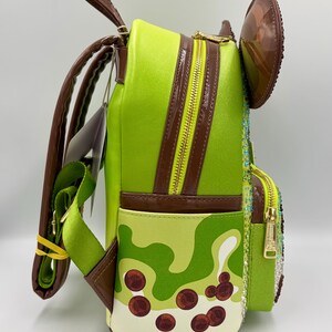 May include: A lime green backpack with brown accents, a gold zipper, and a brown ear-shaped detail. The backpack features a pattern of light and dark green shapes with brown circular details, and a green strap.