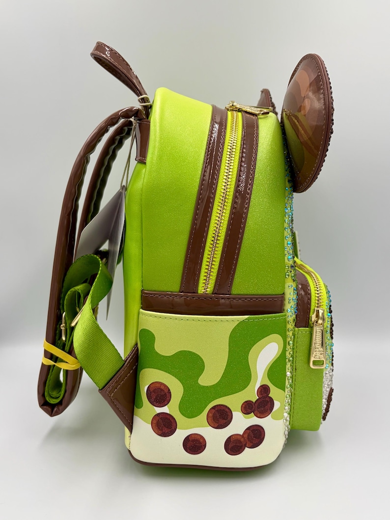 May include: A lime green backpack with brown accents, a gold zipper, and a brown ear-shaped detail. The backpack features a pattern of light and dark green shapes with brown circular details, and a green strap.