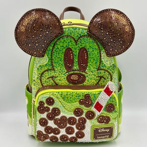 May include: A lime green backpack featuring a Mickey Mouse design with brown ears and a brown handle. The backpack has a zipper closure and is embellished with rhinestones. A red and white striped straw is attached to the front. The Disney Loungefly logo is visible.