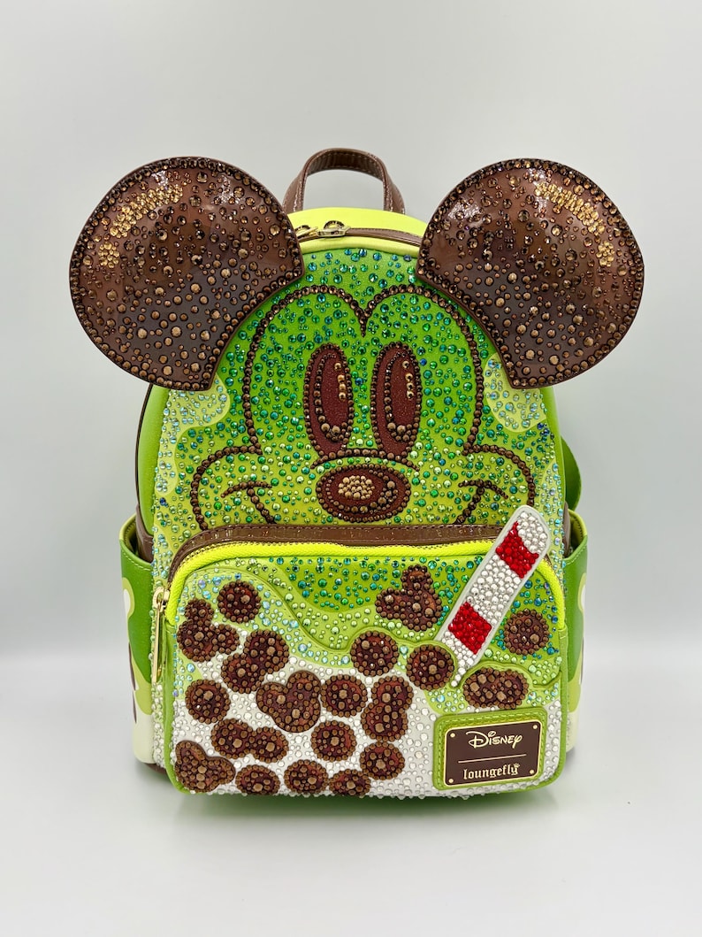 May include: A lime green backpack featuring a Mickey Mouse design with brown ears and a brown handle. The backpack has a zipper closure and is embellished with rhinestones. A red and white striped straw is attached to the front. The Disney Loungefly logo is visible.