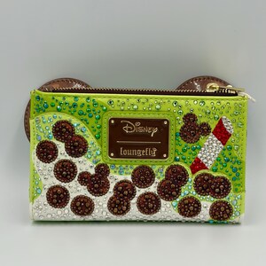 May include: A lime green Disney Loungefly wallet with brown Mickey Mouse ear accents. The wallet features brown, white, and red embellishments, including Mickey Mouse shapes and a candy cane design. The Disney and Loungefly logos are visible.
