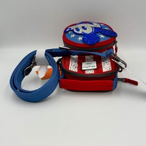 May include: A red, white, and blue crossbody bag featuring a circular top compartment with a blue glittery character applique and a rectangular base with vertical stripes and clear rhinestones. It includes a detachable blue shoulder strap.