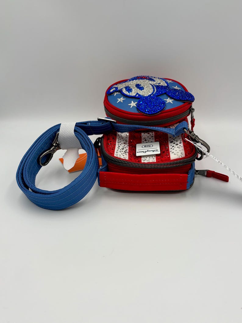 May include: A red, white, and blue crossbody bag featuring a circular top compartment with a blue glittery character applique and a rectangular base with vertical stripes and clear rhinestones. It includes a detachable blue shoulder strap.