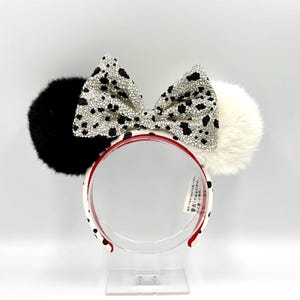 May include: A headband featuring two large fluffy ears, one black and one white, with a central bow covered in silver and black rhinestones. The headband frame is white with black spots and a red inner lining, including a small white tag with text.