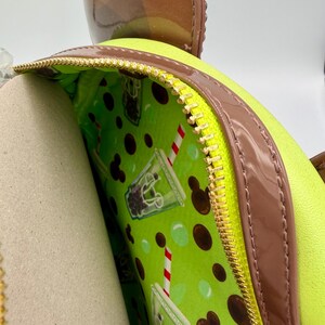 May include: A lime green backpack with a brown trim and a gold zipper. The interior lining is a bright green with a pattern of boba tea drinks and Mickey Mouse silhouettes. A brown bow-shaped accessory is visible at the top.