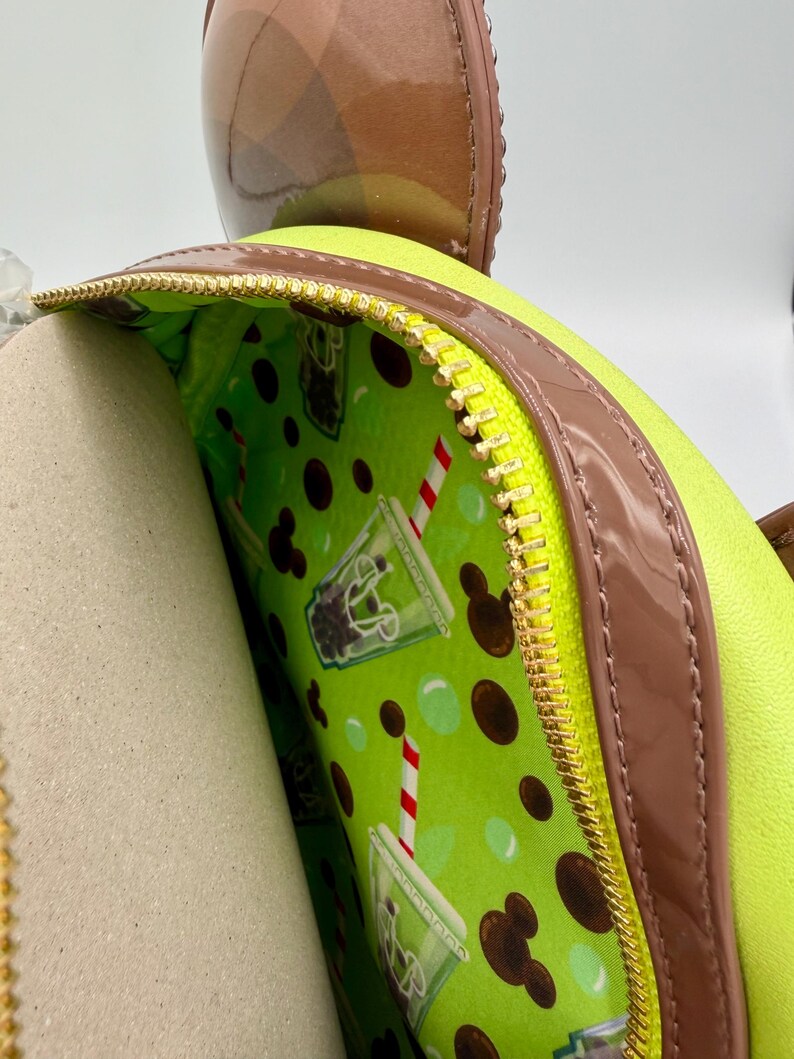 May include: A lime green backpack with a brown trim and a gold zipper. The interior lining is a bright green with a pattern of boba tea drinks and Mickey Mouse silhouettes. A brown bow-shaped accessory is visible at the top.