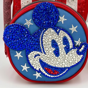 May include: A round blue crossbody bag featuring a crystal-embellished Mickey Mouse face. The design includes blue, white, and red rhinestones forming the character, surrounded by small crystal stars on a blue fabric background with a red border.