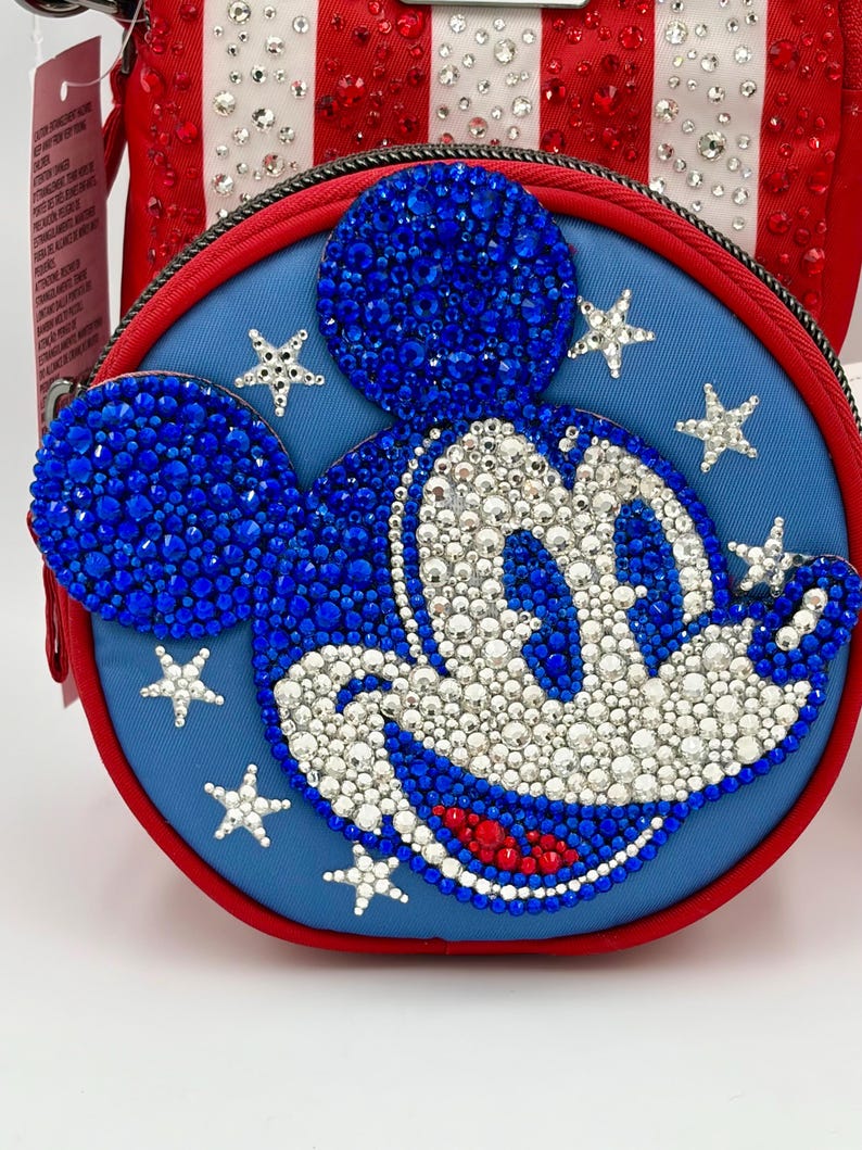 May include: A round blue crossbody bag featuring a crystal-embellished Mickey Mouse face. The design includes blue, white, and red rhinestones forming the character, surrounded by small crystal stars on a blue fabric background with a red border.