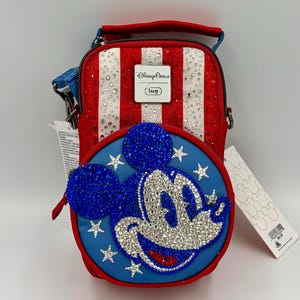 May include: A red, white, and blue crossbody bag featuring a rhinestone-encrusted Mickey Mouse face on a circular front pocket. The bag has vertical stripes, a Disney Parks logo plaque, and a blue strap. It includes attached white product tags.