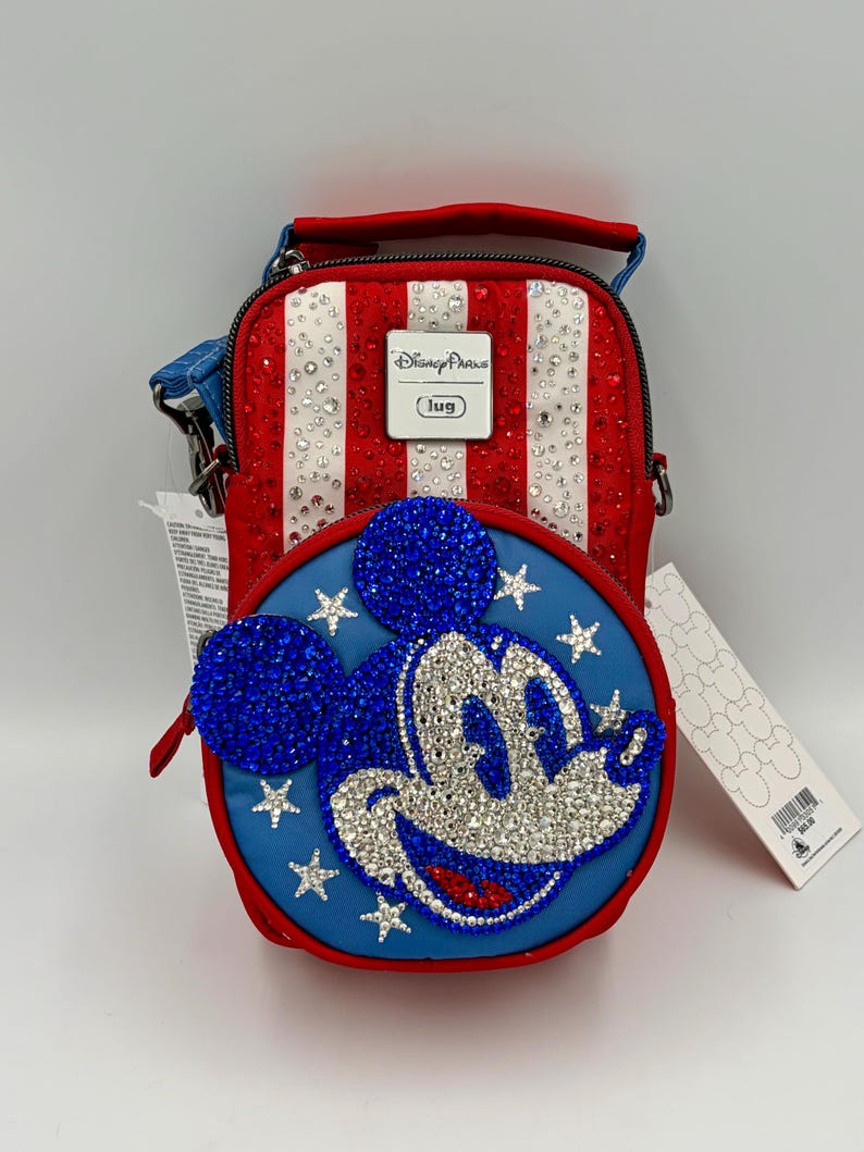 May include: A red, white, and blue crossbody bag featuring a rhinestone-encrusted Mickey Mouse face on a circular front pocket. The bag has vertical stripes, a Disney Parks logo plaque, and a blue strap. It includes attached white product tags.