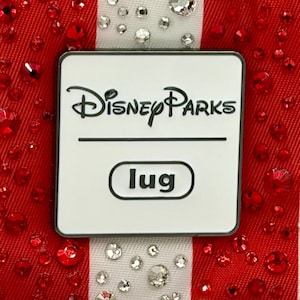 May include: A rectangular white metal plaque with rounded corners featuring the black text Disney Parks and the word lug inside a black-outlined oval. The plaque is centered on a red fabric background with vertical white stripes adorned with clear crystals.